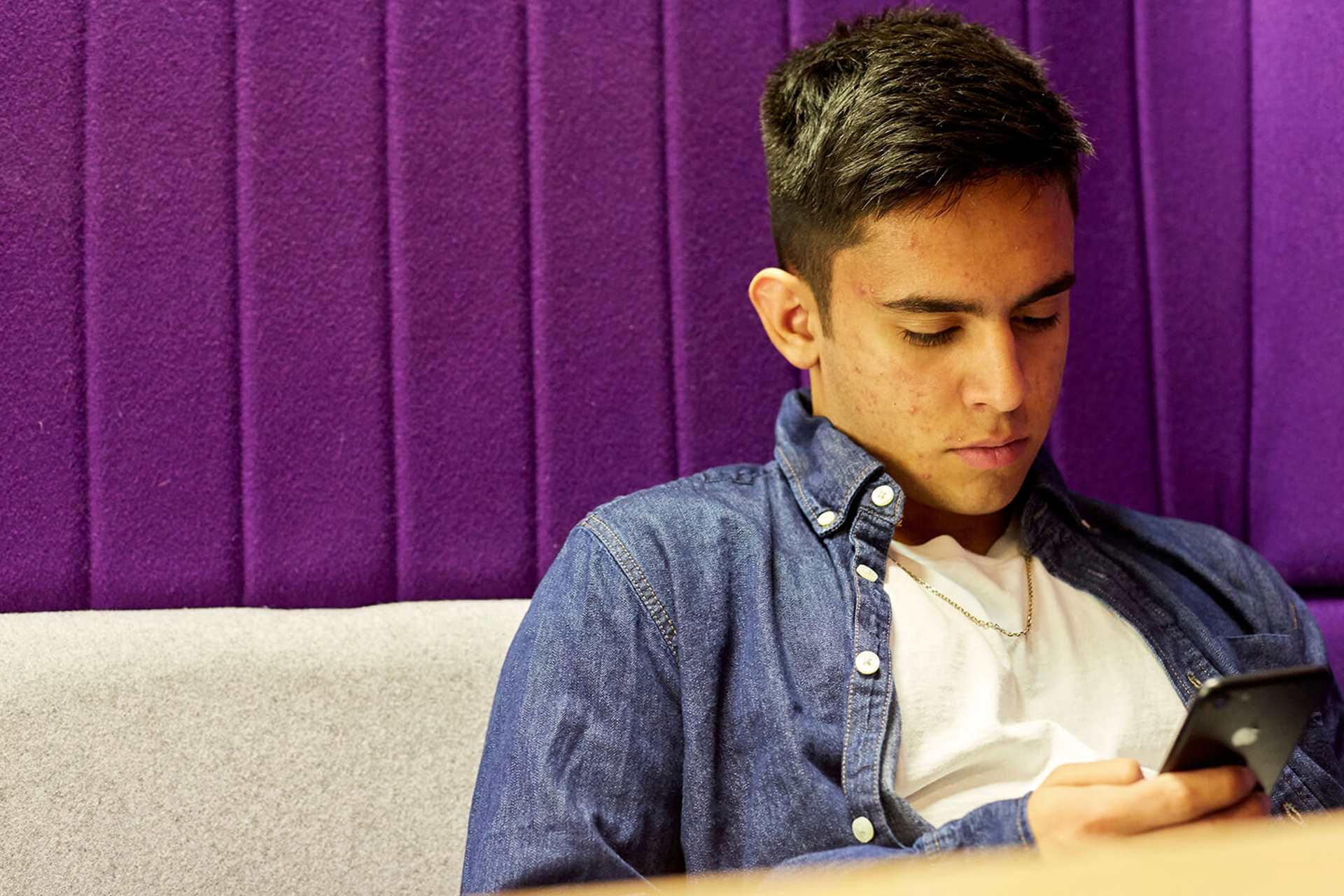 A boy in a blue denim shirt looks at his phone while sitting down against a grey sofa with a purple wall.