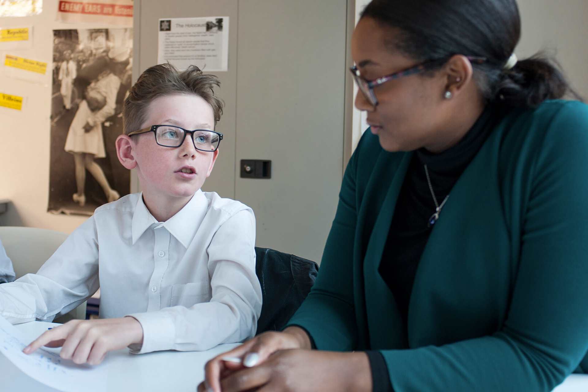a boy wearing school uniform and glasses sits on his desk holding a paper asking help from his teacher sitting beside him in the classroom