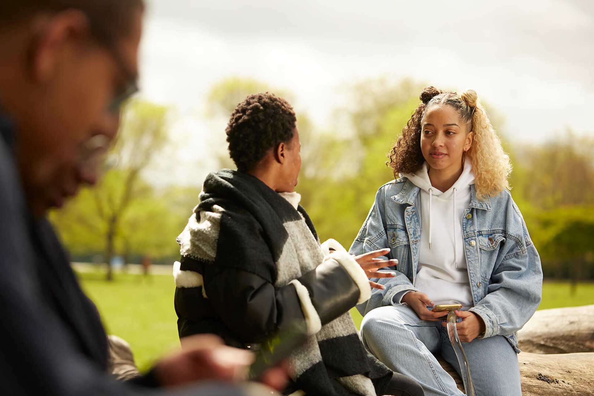 Two girls sit and chat on a fallen tree trunk in a park.