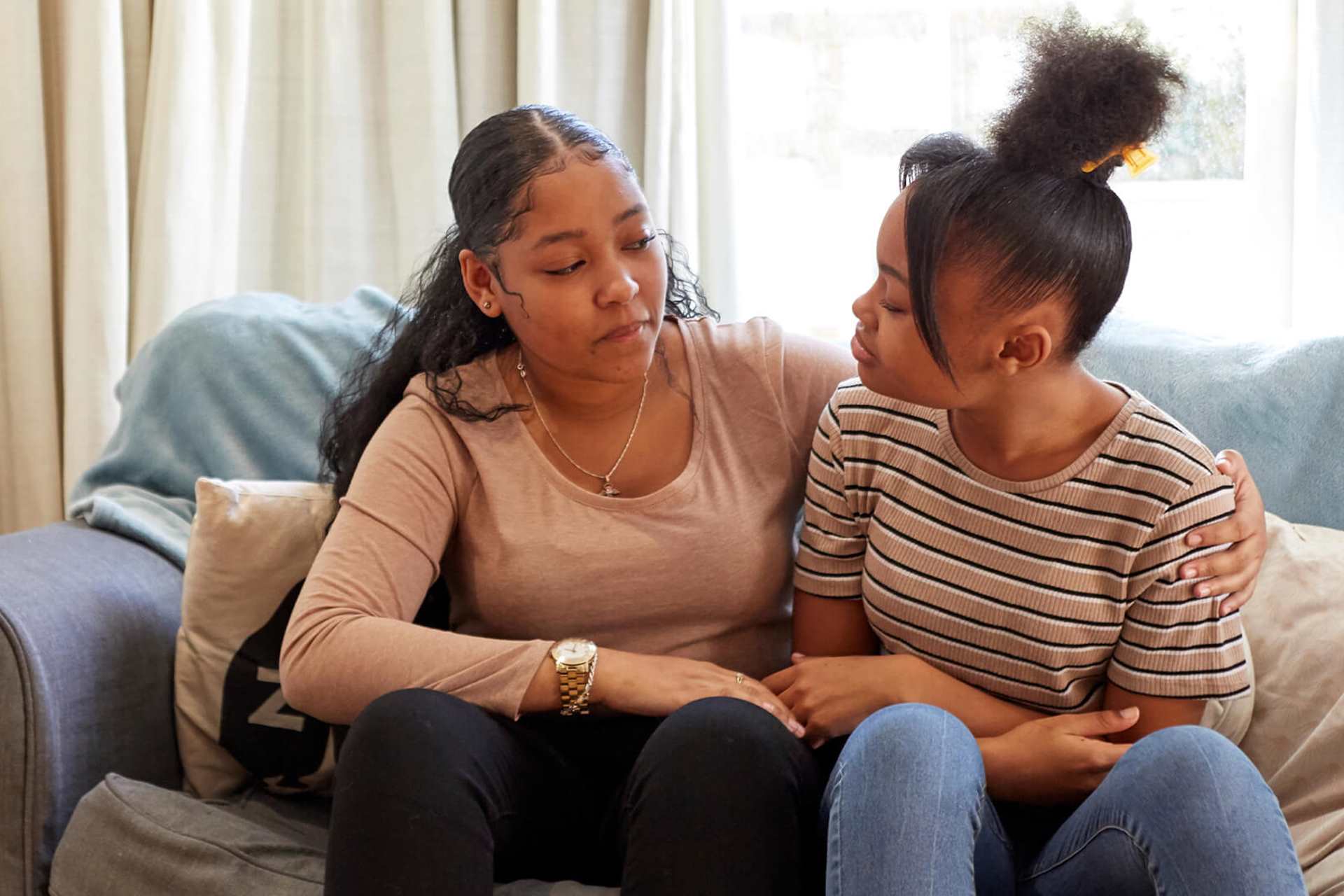 Two young people sit on a sofa with the person on the left putting an arm around the other. They both are looking at each other while talking.