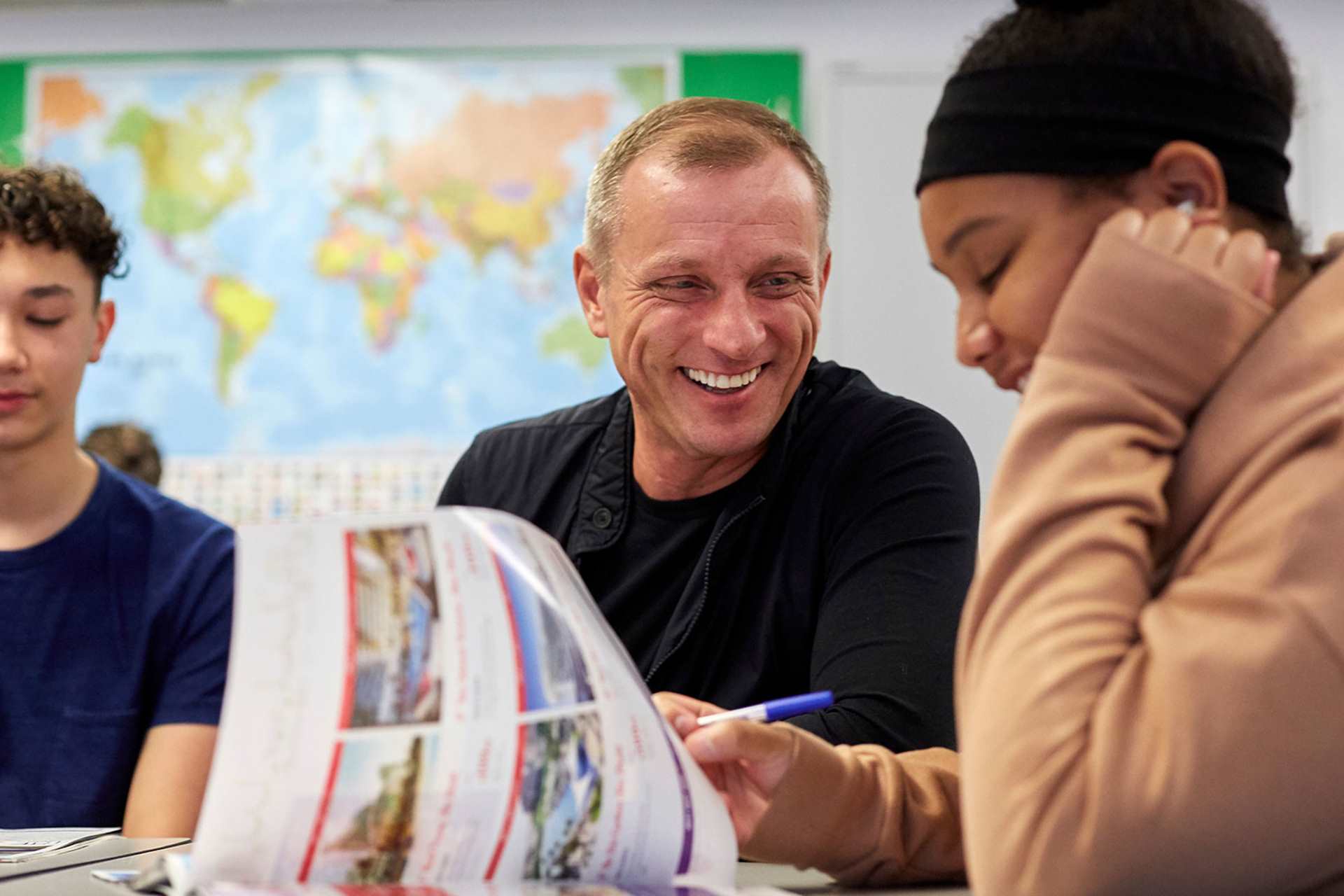 a teacher is laughing while he sits in between two of his students who are also laughing inside the classroom