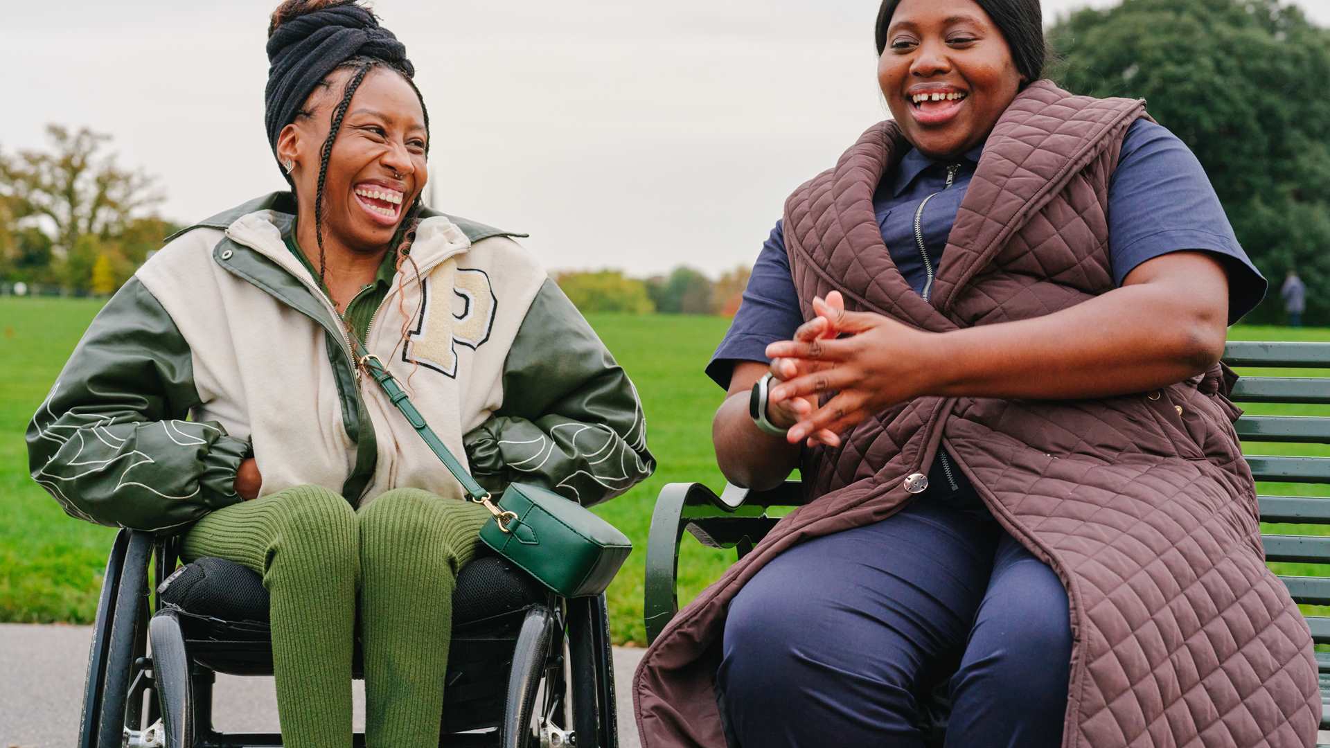 A young Black woman in a wheelchair and an older Black woman sitting on a bench in the park. They are laughing together.