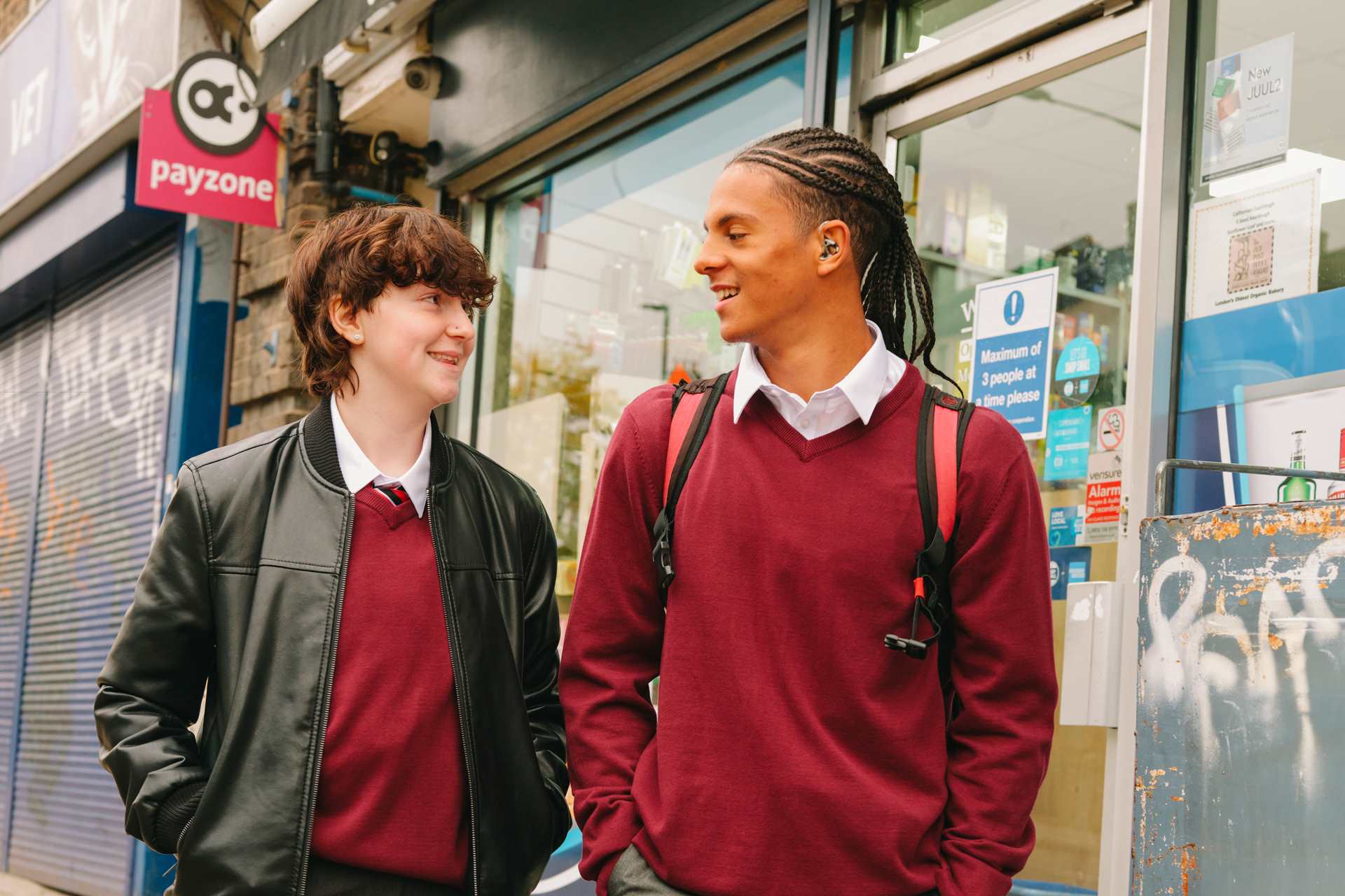A Black teenage boy wearing a hearing aid speaking to a white non-binary teenager. They are walking on the street outside a shop. Both people are smiling.