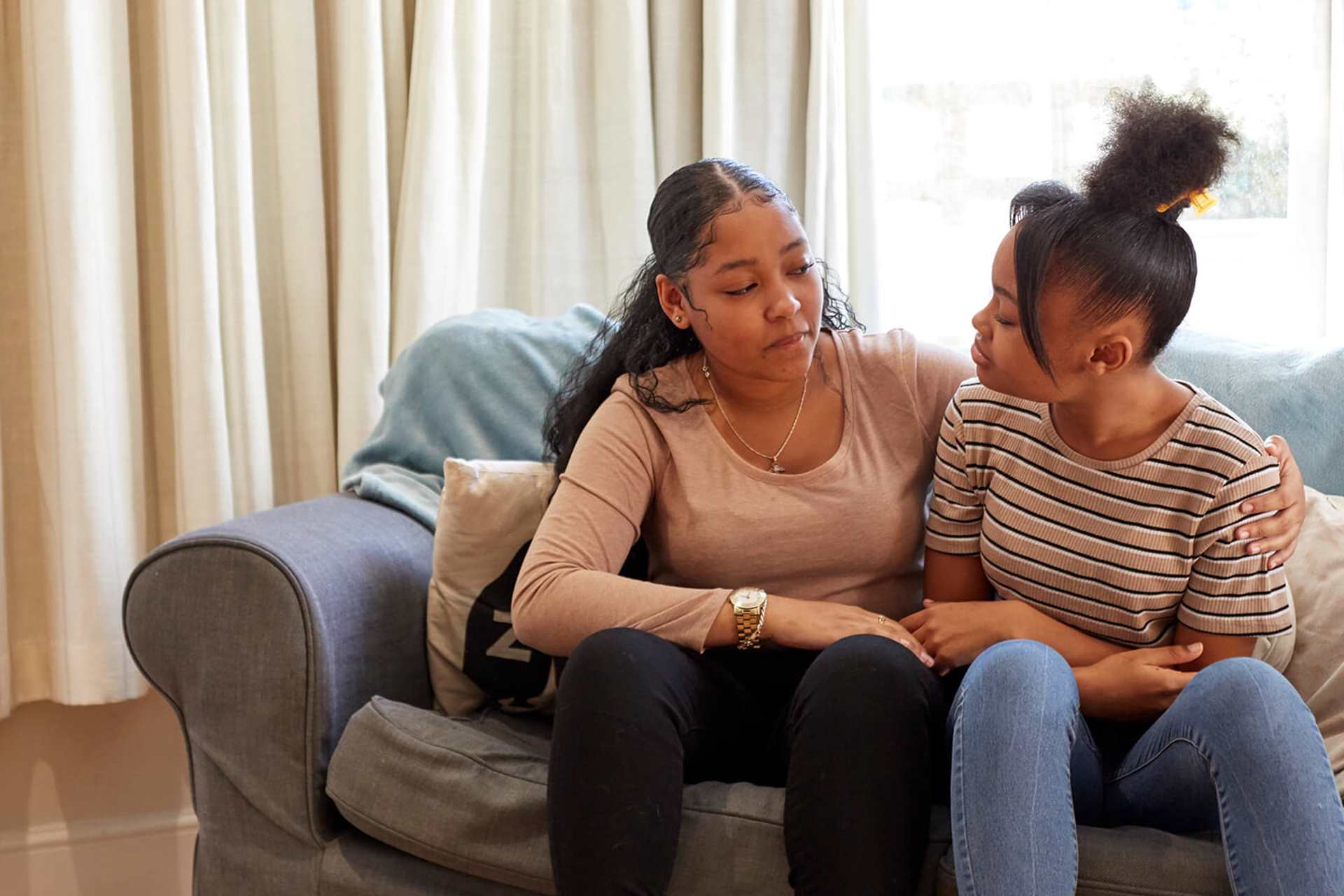 Two young people are sitting on a blue sofa facing each other. The person on the left has their arm around the person on the right.