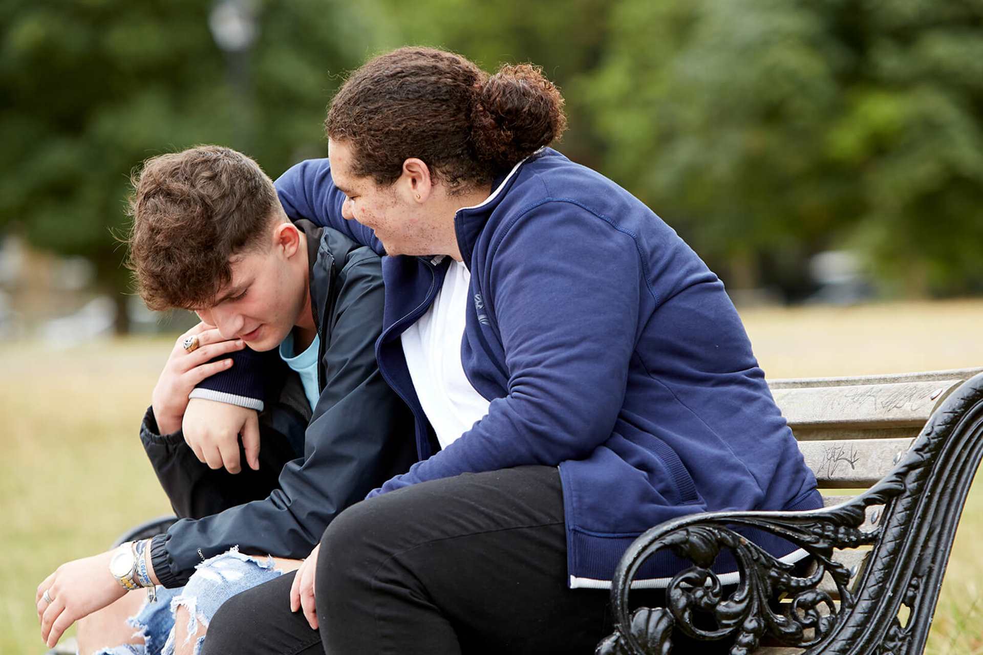 Two young people sit on a bench in a park. The person on the right has his arm around the other young person. The young person on the left is holding the other persons arm while looking down at the floor.