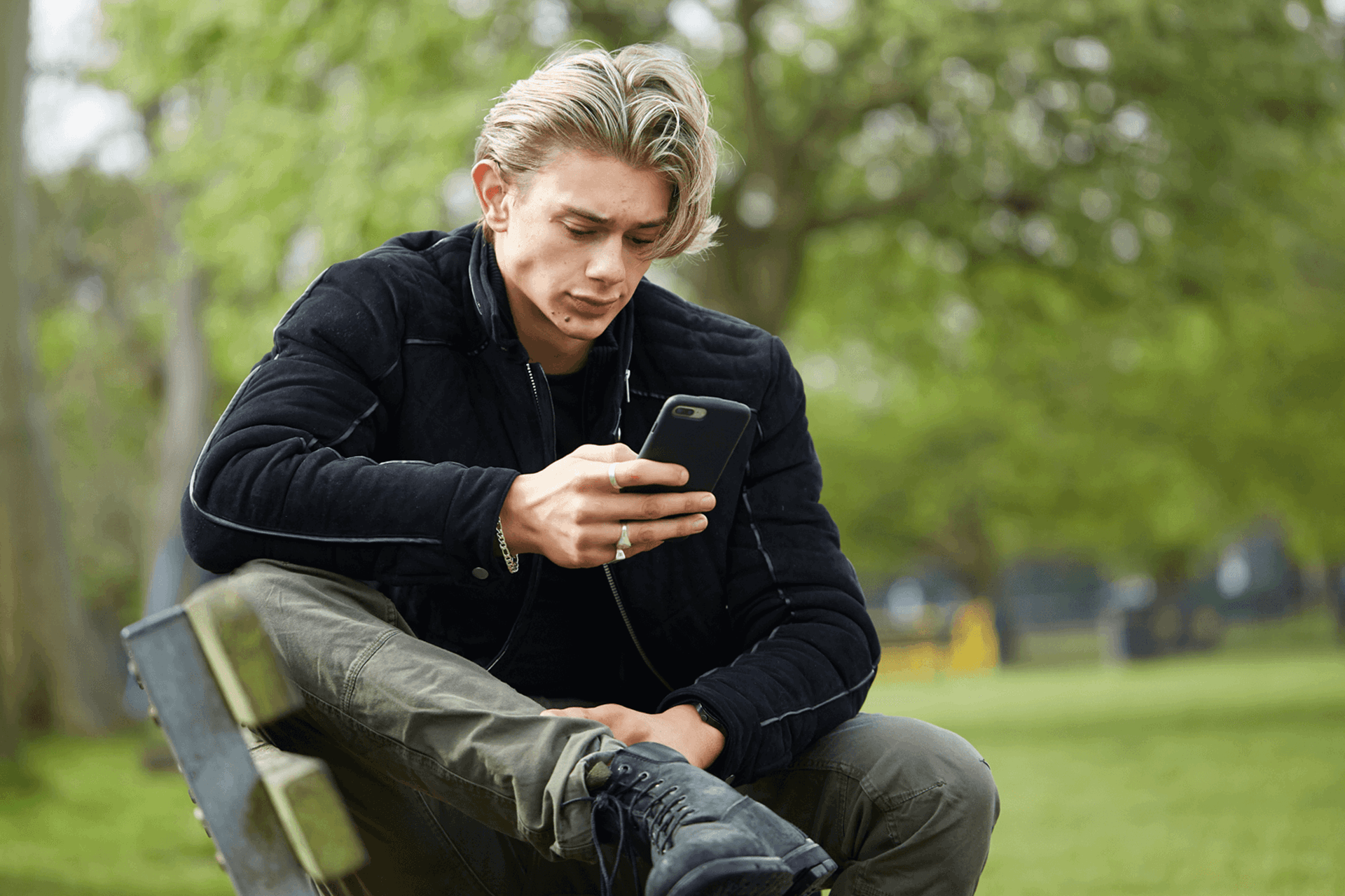 A young man wearing a black jacket sits on a park bench. He is looking at his phone with a worried expression.