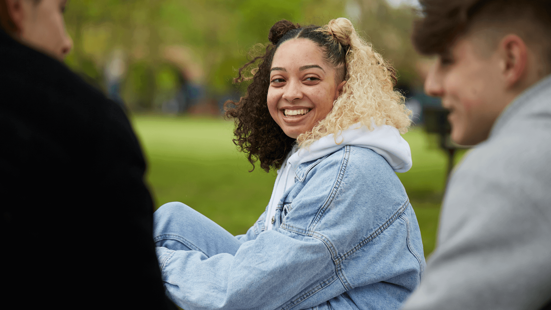 A young woman with curly hair wearing a blue denim jacket is smiling and talking with two young men in the park.
