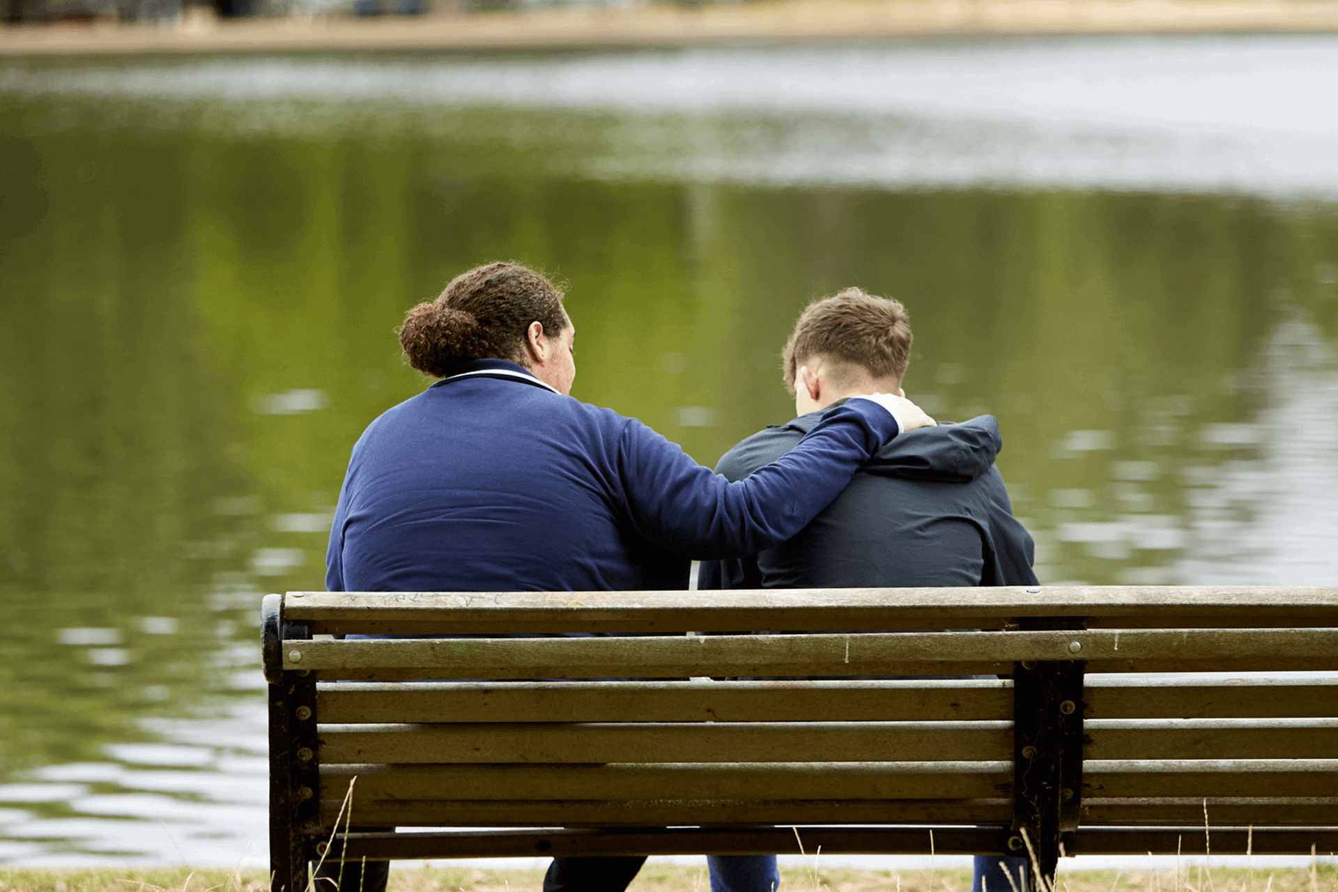Two young people sitting on a bench in a park, one has their arm around the other.