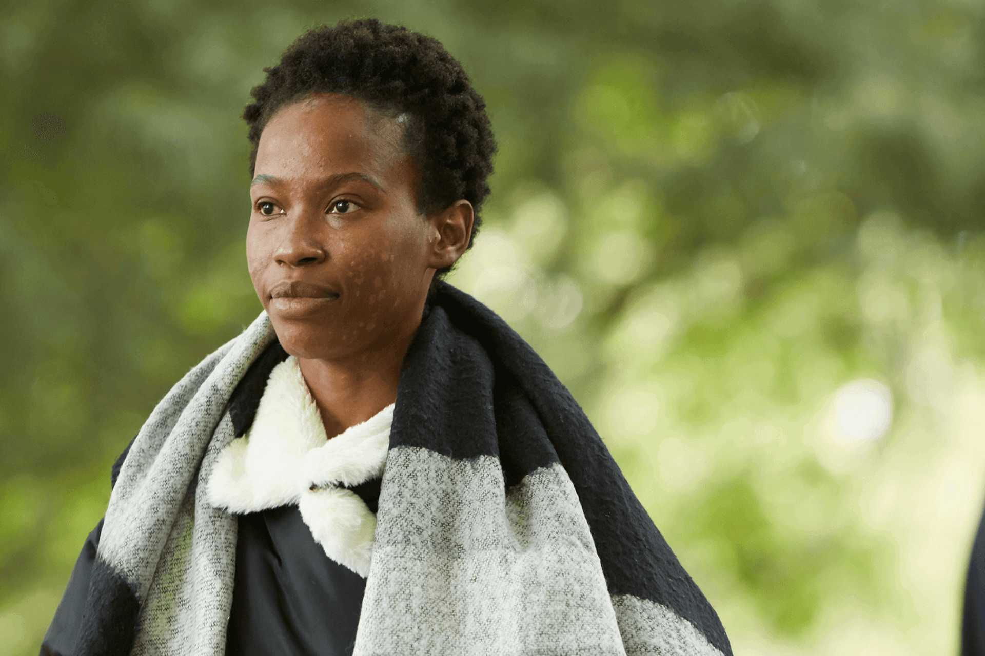 A young woman wearing a grey and black scarf walking through a park.