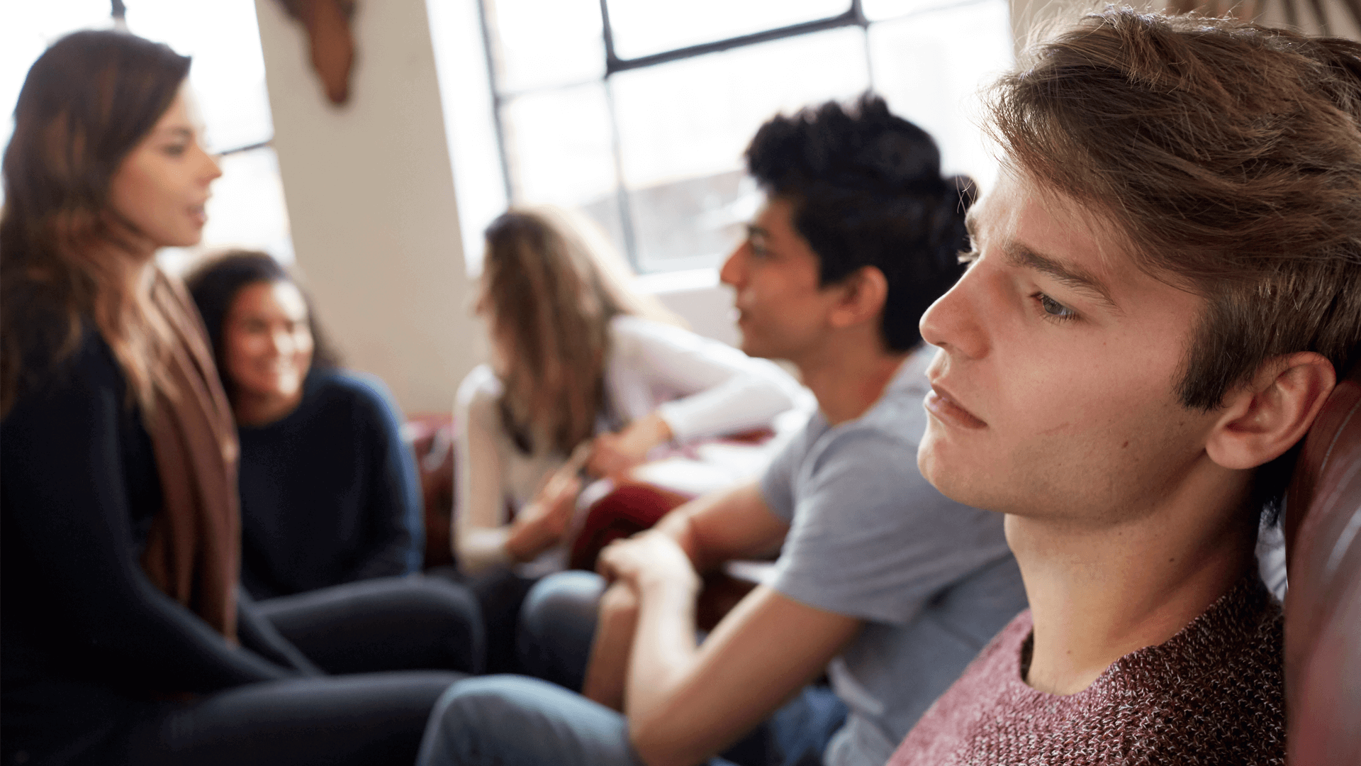 A young person lost in thought while sitting with their group of friends who are talking together.
