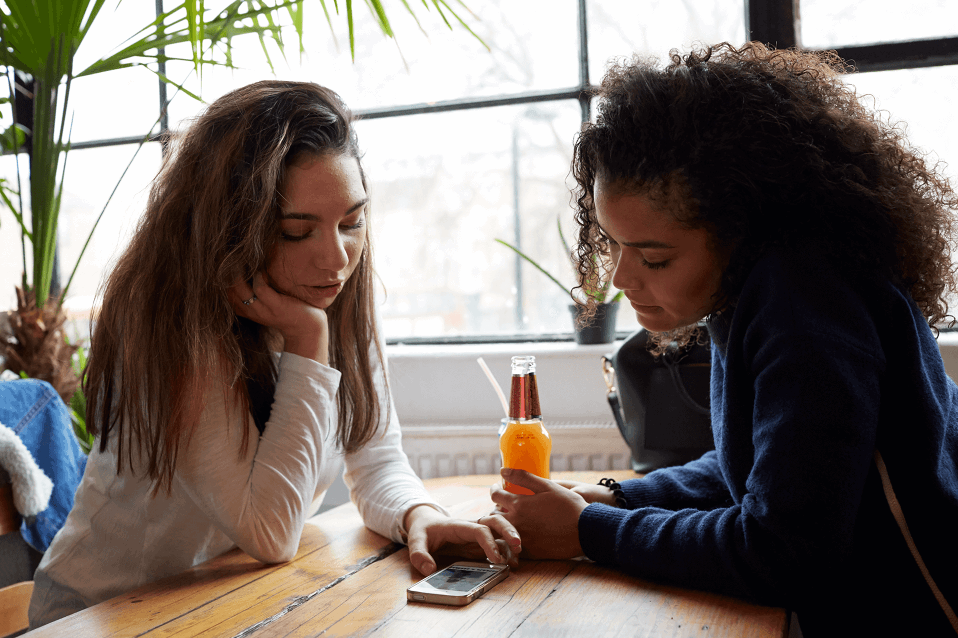 Two young women sit opposite each other at a wooden table in a restaurant. They are both looking at something on one of their smart phones, which is lying flat on the table between them,