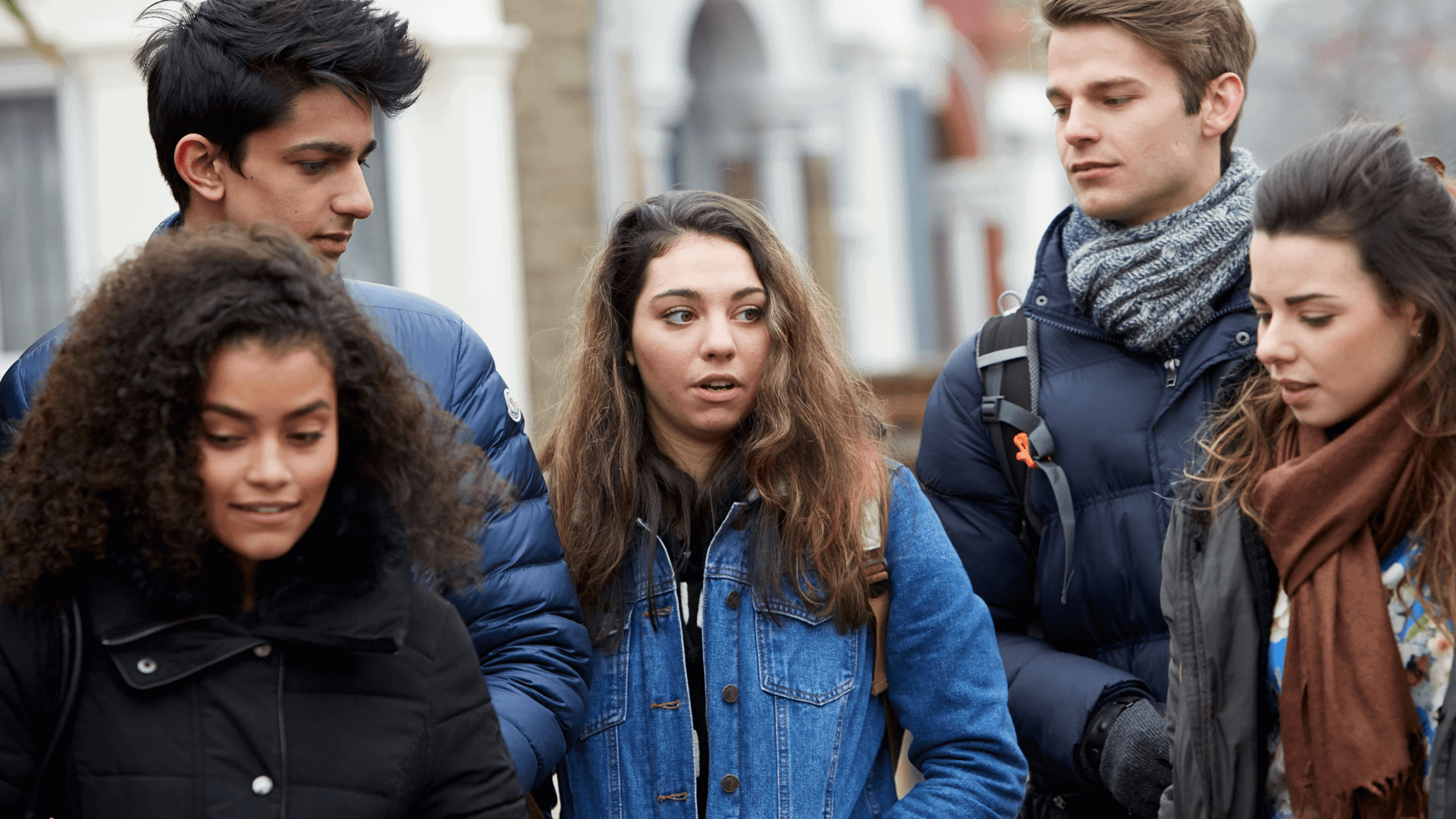 Five young people wearing coats, jackets and backpacks. They are talking as they walk down a street.