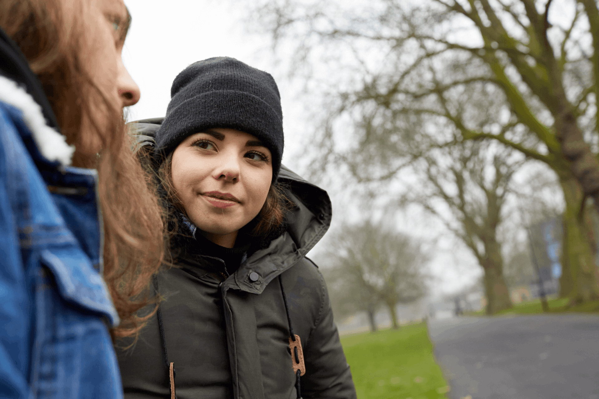 A girl in her winter coat is looking and smiling at her friend as they are talking while sitting on a park bench.