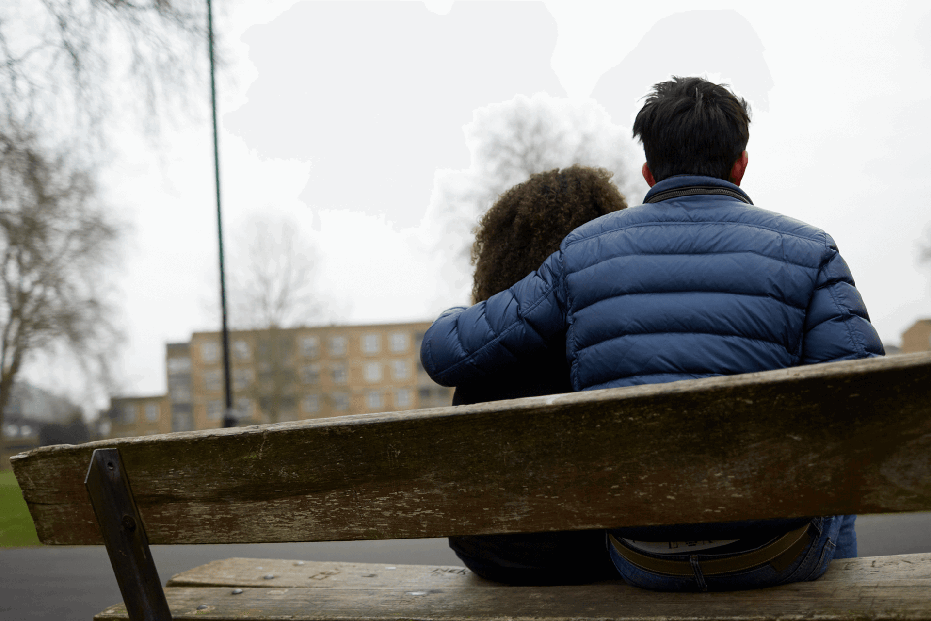 Two young people sitting together on a bench. One has their arm around the other.