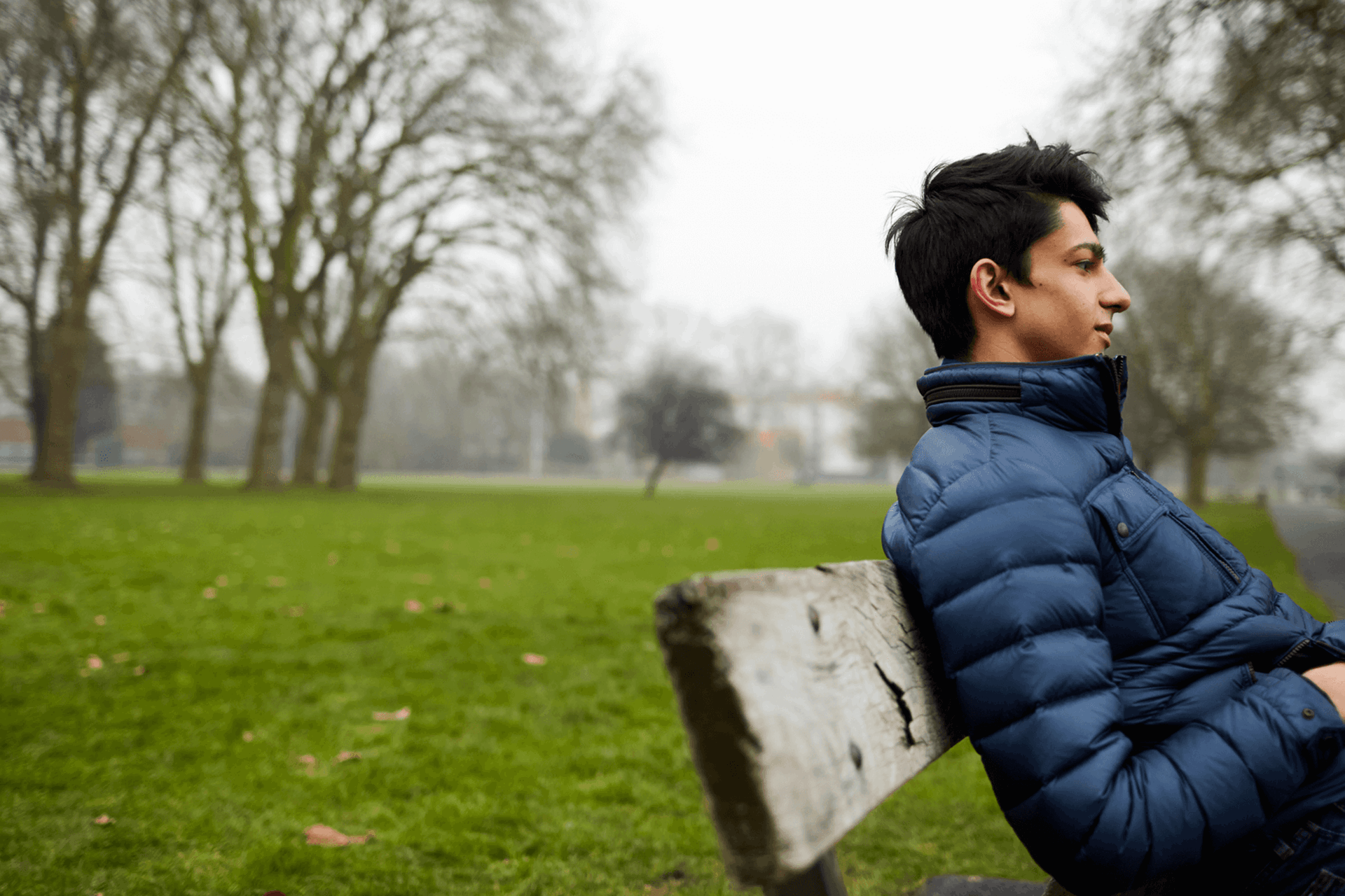 A boy sits on a park bench wearing a blue puffer jacket with his hands in his pockets. He is looking straight ahead as if deep in thought.