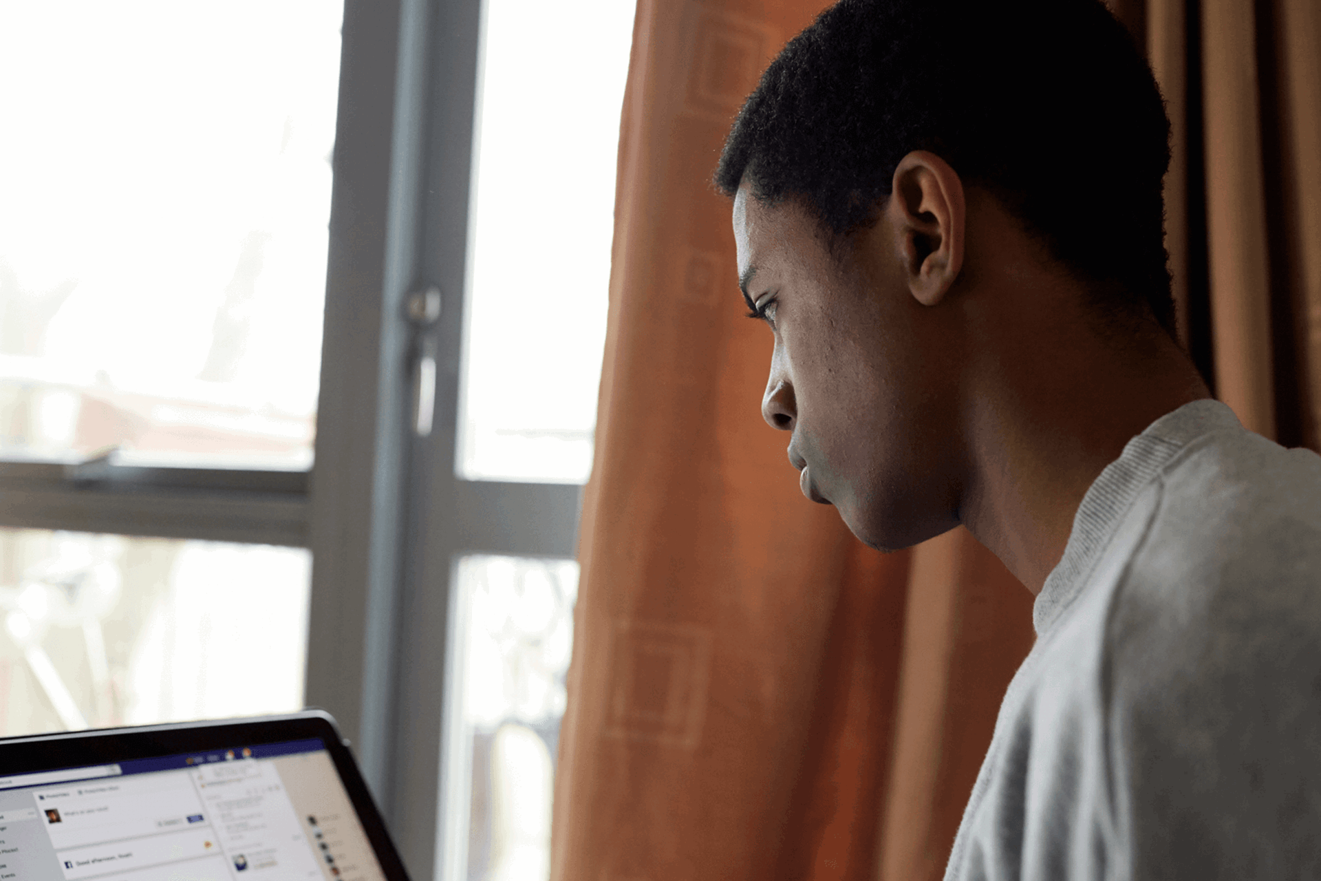 A boy wearing a grey t-shirt sits beside a window while using Facebook on his laptop.