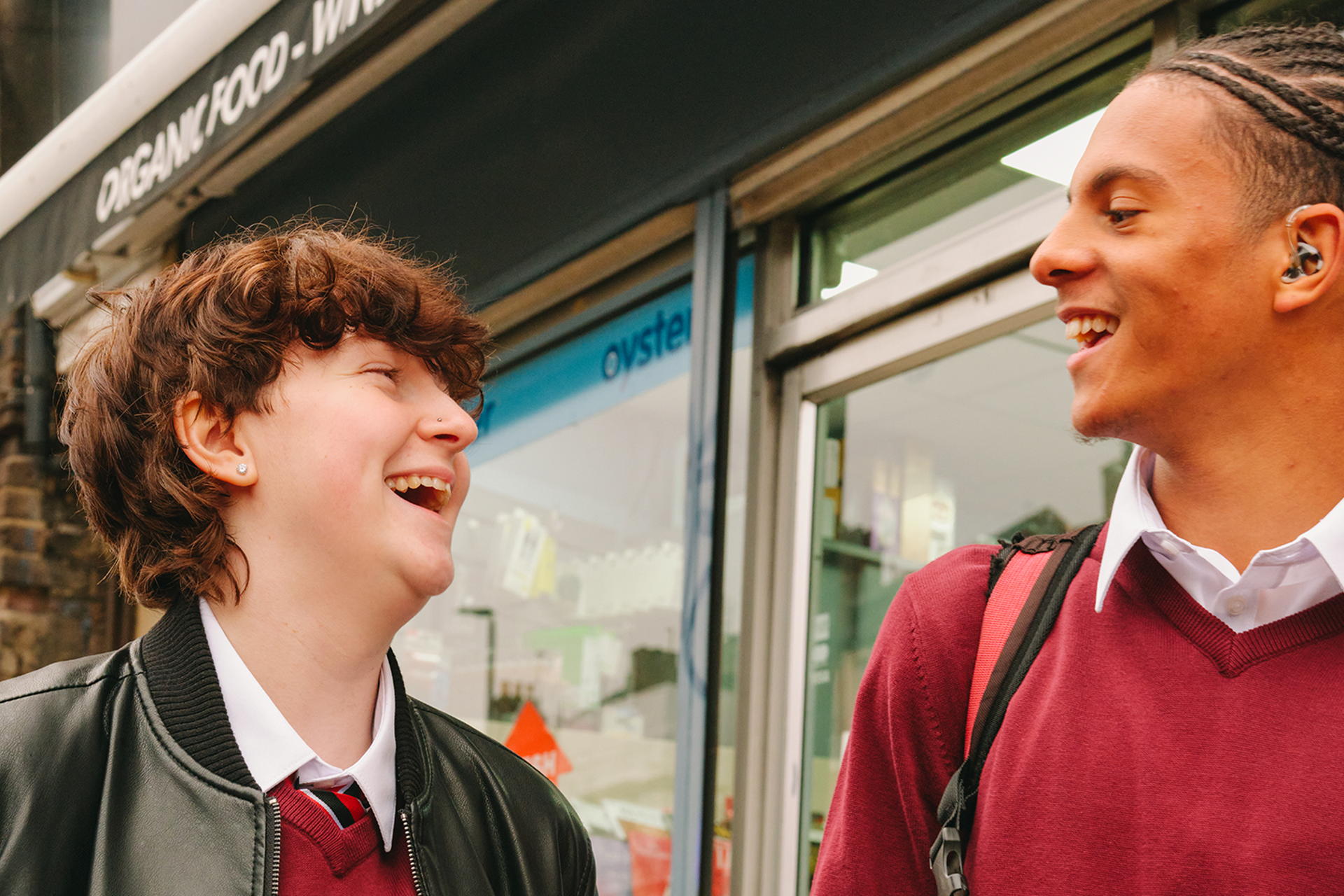 A Black teenage boy wearing a hearing aid laughing with a white non-binary teenager outside the shops.