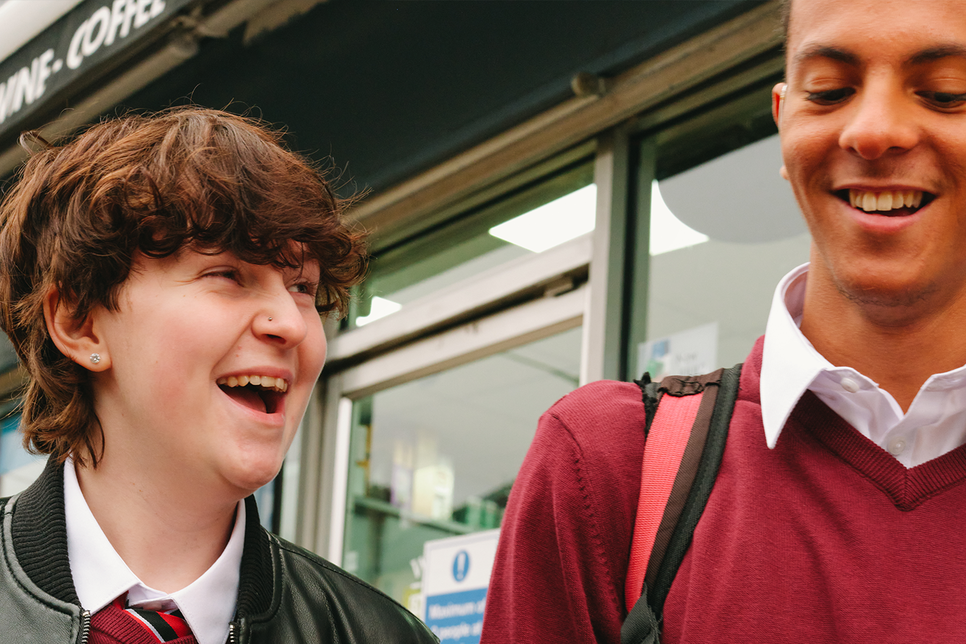 A Black teenage boy wearing a hearing aid laughing with a white non-binary teenager outside the shops.