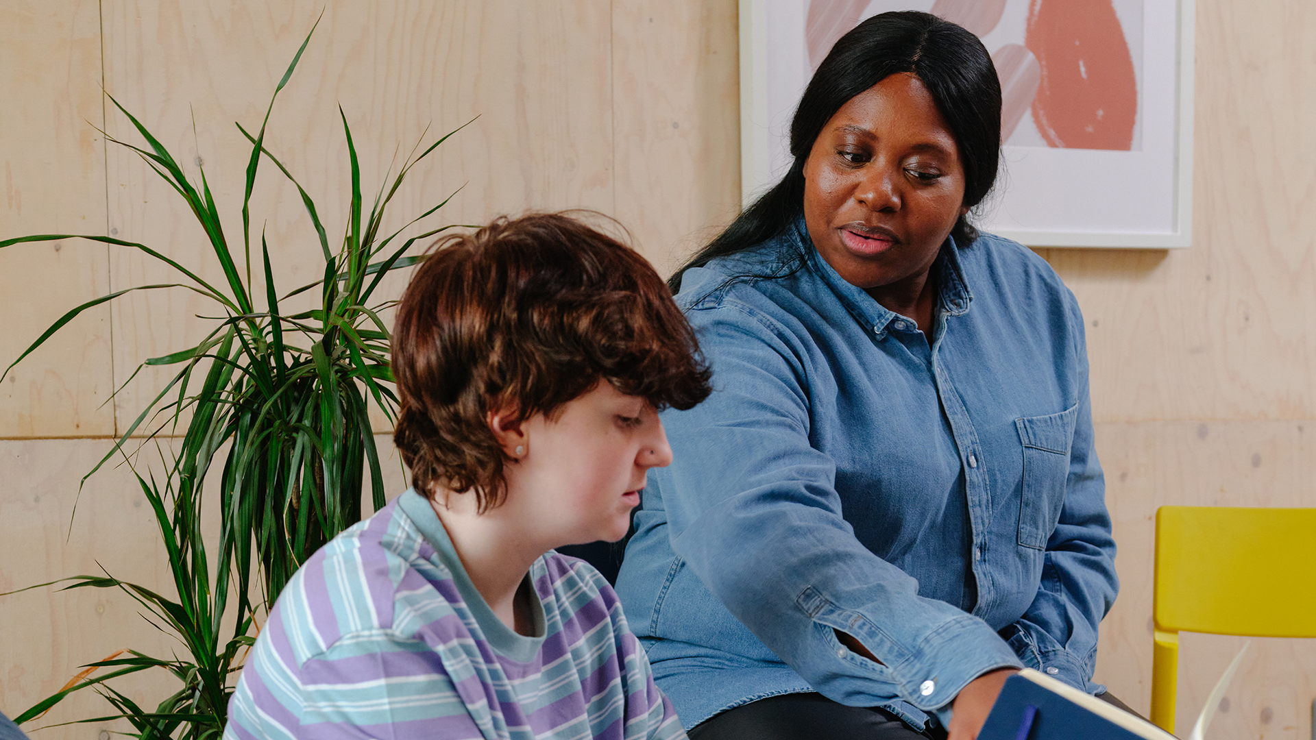 A white non-binary teenager reading with an older Black woman in a professional setting.